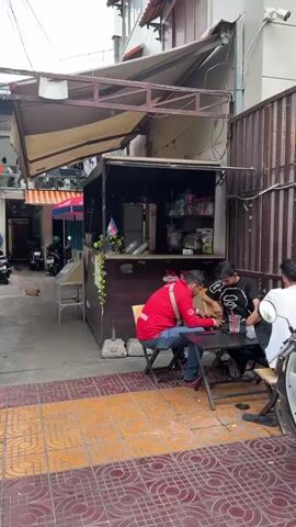 The video shows an alleyway scene with a small food stall on the right. Two men are seated at a small table in front of the stall. One man is wearing a red jacket and the other is wearing a black shirt. Several motorcycles are parked along the left side of the alley. An orange cat is lying on the ground in the middle of the alley. A white plastic bag is on the ground to the far left of the frame.
