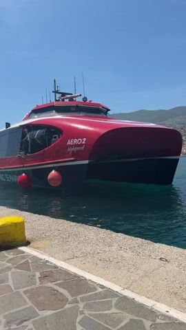 A large red and black high-speed catamaran ferry, named "AERO 2 Highspeed," is featured prominently in the video. The ferry is either docked or preparing to depart/arrive, with its engines making a noticeable sound and churning the water at its stern. A person is visible on the deck near the front, possibly attending to mooring lines.

The ferry has "HELLENIC SEAWAYS" written in white lettering along its side and several red buoys hanging from its port side. The scene is set on a clear, sunny day, indicated by the bright blue sky. In the foreground, a stone dock with a yellow bollard is visible. The water is a deep blue-green. Rolling hills, partially covered in greenery, form the backdrop, suggesting a coastal or island location. The overall atmosphere is bright and calm, typical of a Mediterranean port.