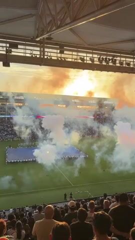 The video shows a wide shot of a soccer stadium during a pre-game ceremony. Smoke fills the air on the field, obscuring many of the players who are lined up. Spectators are seated in the foreground, facing the field. The sun is setting in the background, casting a warm glow over the scene. A large Jumbotron screen displays an image of an American flag and a person singing. The stadium appears to be outdoors with a metal roof structure overhead.