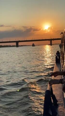 This video captures a serene sunset over the Ob River in Novosibirsk, Russia. The sun, a bright orb, hangs low in the sky, casting a warm, golden glow across the water. The sky is painted with soft clouds tinged with orange and pink hues. A prominent bridge, likely the Bugrinsky Bridge given its distinctive arch, spans the river in the distance. The bridge is illuminated by the setting sun, with its lights beginning to twinkle.

A boat, on which the video is being filmed, is moving along the river. The camera is positioned to show the wake of the boat rippling on the water's surface, creating shimmering reflections of the sunlight. In the foreground, the edge of the boat is visible, along with two individuals' hands resting on the railing. Their relaxed posture and the peaceful ambiance suggest a leisurely moment. A cargo ship is seen in the middle distance, adding a touch of industrial activity to the natural beauty of the scene.

The time of day is clearly sunset, indicated by the low sun and the warm, diffused light. The weather appears to be pleasant, with no visible rain or strong winds. The scene evokes a sense of tranquility and awe, a common feeling experienced when witnessing a beautiful sunset over a significant body of water. The overall mood is peaceful and contemplative, offering a glimpse into a tranquil evening by the Ob River. There is no visible text in the video.
