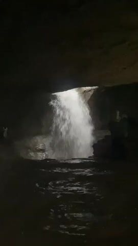 The video shows a cave with a waterfall flowing into a pool of water. The cave is dark, with the waterfall being the main source of light. There are people visible in the cave, some standing near the waterfall and others on a ledge above it. The water is clear and appears to be moving swiftly. The rocks in the cave are wet and reflective. The video pans from the waterfall to the surrounding cave walls and then to a person standing on the ledge.