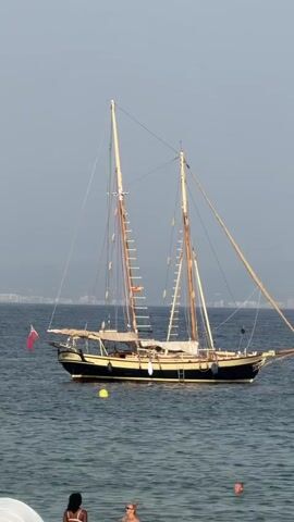 A medium-sized sailing yacht is anchored in calm, clear blue water near a beach. The yacht has two masts and appears to be old-fashioned. A red and white flag flies from the stern. In the foreground, a few people are in the water, swimming and relaxing. The sky is clear and pale blue, suggesting a sunny day. The overall scene is peaceful and idyllic.