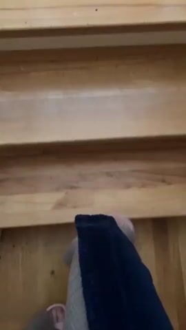 The video shows a first-person perspective of someone walking down wooden stairs. The subject is wearing a dark blue robe and light-colored slippers. The stairs appear to be indoors, likely in a home. The lighting suggests it is daytime, but the weather is not discernible. There are no visible signs of text in the video. The activity is a simple act of descending stairs, and the overall mood seems casual and relaxed. The wooden texture of the stairs and the attire of the subject contribute to a homey and comfortable atmosphere.
