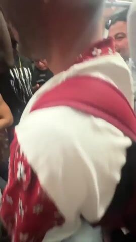 The video shows a group of people inside a crowded public transport vehicle, likely a train or bus, as indicated by the overhead handrails and the close proximity of passengers. The focus is on a young man in the foreground wearing a white t-shirt with red bandana-style trim around the neckline and sleeves. He has tattoos on his arms and is wearing a gold earring. He appears to be speaking or shouting directly into the camera, with his mouth open wide. In the background, other passengers are visible, including a man with numerous tattoos on his arm and a man wearing a red kufi cap and red shorts. The lighting is dim and appears to be artificial. The overall atmosphere suggests a lively or energetic event, possibly a celebration or a journey with friends.