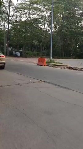 The video shows an outdoor scene on a paved road. The camera is in motion, moving from right to left. In the foreground, a person wearing a yellow hoodie and dark shorts is riding a motorcycle. The motorcycle is black and appears to be a scooter. The person's face is not clearly visible due to motion blur and the angle. In the background, there are trees and a light pole. Further down the road, another motorcycle carrying a large red box is visible, ridden by a person wearing a light-colored shirt and a cap. The red box on the motorcycle appears to contain items, possibly for sale or delivery. There are also orange traffic cones visible on the side of the road. Further in the distance, there are some structures and more trees. The lighting suggests it is daytime, possibly late afternoon or early morning, with soft sunlight filtering through the trees. The road surface appears to be asphalt with some visible cracks and markings. The overall scene depicts a street environment with traffic and vegetation.