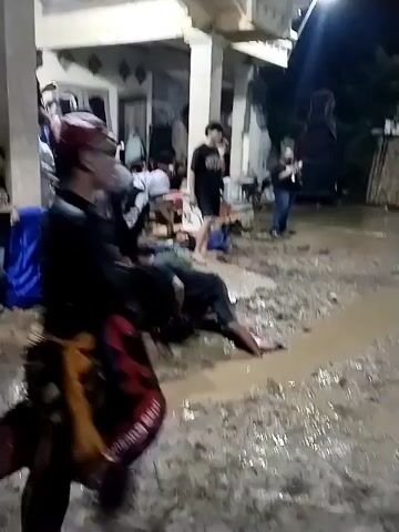 The video captures an outdoor night scene in Jenggrik, Indonesia, featuring a gathering of people. The ground is heavily muddy with significant puddles and a stream of muddy water. In the center, a person lies on the muddy ground, seemingly in a trance or an altered state, with their lower body partially submerged in a muddy puddle. Two women, one wearing a hijab, are actively attending to this person, bending over them near a small wooden bench. Other individuals are standing around, observing the scene. In the foreground, a person wearing a black top and a red and black patterned lower garment, possibly a performer, walks away from the camera. Artificial lights illuminate the area, and a building with pillars and an arched entrance can be seen in the background, along with large speakers. The setting suggests a community event or traditional performance taking place under challenging ground conditions.