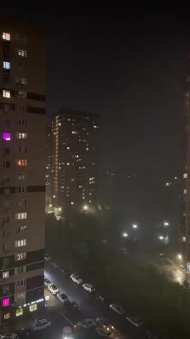 The video shows an urban nighttime view between tall buildings, captured during a thunderstorm.  Rain is falling, and visible flashes of lightning briefly illuminate the scene. Cars are parked along the street below.  Building lights are visible in the surrounding structures, with one apartment showing a distinct purple hue.  Trees can be seen between buildings and along the street.