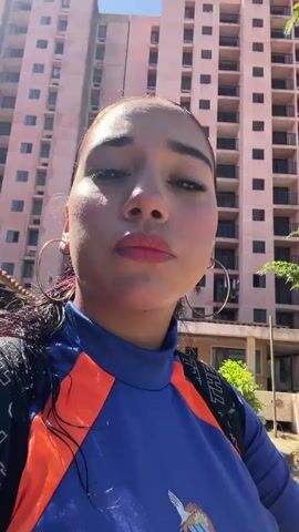 The video shows a woman in a blue and orange long-sleeved shirt posing for a selfie. She is outdoors, with a building in the background and greenery around her. She is wearing hoop earrings and has her hair pulled back. The video is brief and does not show any specific actions or interactions.