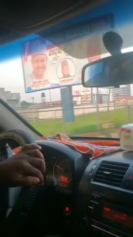 The video appears to be filmed from inside a moving vehicle, looking through the windshield. The driver's hands are visible on the steering wheel, and an orange cloth is draped over the dashboard. In the foreground, a box of tissues sits on the dashboard. Through the windshield, a street scene unfolds. Several vehicles, including a yellow auto-rickshaw, are on the road. Buildings and billboards are visible in the background. The sky is overcast, and the overall lighting suggests it is daytime. People are seen on the sidewalks and near the road. The footage is shaky, consistent with being recorded from a moving vehicle.