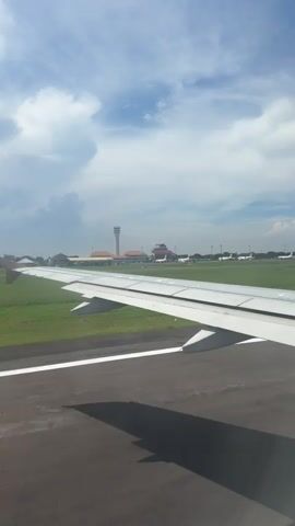 A video taken from an airplane window shows the aircraft taxiing on a runway at an airport. The white wing of the airplane is prominent in the foreground, with its flaps adjusting. The dark asphalt runway, marked with white and yellow lines, is visible below the wing, with a shadow of the wing cast upon it. Green grass borders the runway. In the distance, an airport control tower and several other parked airplanes are visible under a bright blue sky with scattered white clouds. The scene depicts a sunny day at an airport in Sedati Agung, Indonesia.