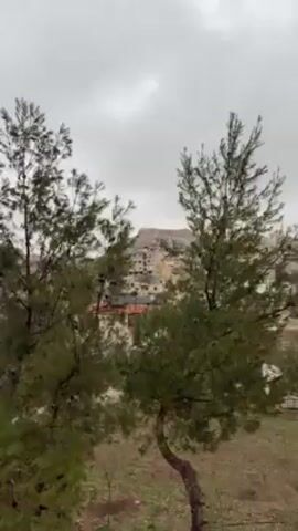 The video presents a static, medium-shot view of a landscape under an overcast sky. Two evergreen trees with lush green foliage stand prominently in the foreground, slightly obscuring the view of a town nestled among hills and rocky terrain. Buildings of varying heights and colors, primarily beige and white with red roofs, dot the cityscape. A mountain range looms in the background, its rocky texture visible despite the diffused light. The weather appears gloomy due to the heavy cloud cover. In the soundtrack, sounds of traffic noises can be heard.