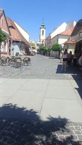 The video shows a cobblestone street in a European town on a sunny day. Buildings with terracotta roofs line both sides of the street. On the left, there are cafe tables and chairs set up outside a building. On the right, there are shops with awnings and goods displayed outside. Several people are walking down the street, some appearing to be tourists. A church spire is visible in the distance at the end of the street. The camera is moving forward, following the street.