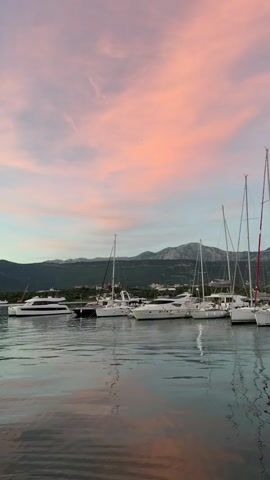 The video displays a panoramic view of a marina at sunset. In the foreground, the calm water reflects the pastel hues of the sky and the silhouettes of mountains in the distance. Several yachts and sailboats are moored in the marina, their masts reaching towards the colorful sky. The sky is painted with shades of pink, orange, and blue, with wisps of clouds adding texture to the scene. The overall atmosphere is serene and picturesque, capturing the tranquil beauty of the coastal landscape as day transitions into night.