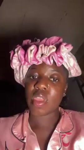 A dark-skinned young woman with a lip piercing wearing pink pajamas and a pink and white floral bouffant cap on her head is visible from the chest up in this video. The woman is looking directly at the camera, she opens her mouth slightly and puts a small amount of white cream or foam onto her upper lip. The background is dark, and the top of a wooden ladder is visible in the right upper corner of the frame. The lighting is poor.