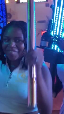The video shows a young Black woman with braided hair and a nose ring, wearing a white crop top, dancing at what appears to be a party or club. She is holding a metallic pole with colorful LED lights attached to it. The environment is dimly lit with vibrant neon lights casting purple, green, and blue hues on the woman and the surroundings. In the background, there are bottles, possibly champagne or liquor, and decorative elements that suggest a festive atmosphere. The woman is smiling and moving her body to the music, interacting with the light-up pole. The camera is close-up on her, capturing her movements and expressions.