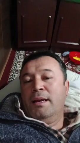 The video starts with a man lying down looking at the camera. The camera quickly pans to a young girl with braided pigtails smiling at the camera. The background includes a patterned rug, a white marbled wall, and a ceiling with a grid-like design and a light.