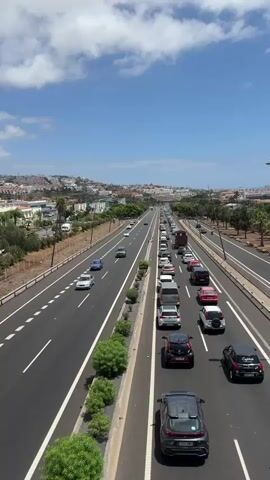 A high-angle, vertical video shows a multi-lane highway under a bright blue sky with scattered white clouds. The highway is congested with traffic in the right lanes, with cars bumper-to-bumper. The left lanes are moving more freely. On the left side of the highway, there are some trees and bushes, and beyond that, a hillside with buildings and more vegetation. On the right side of the highway, there are more trees and a sound barrier. The traffic is moving away from the camera.