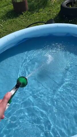 A hand holds a green and black garden hose nozzle and sprays water into a bright blue inflatable pool filled with water. The water from the hose creates a powerful jet that splashes into the pool, causing ripples and disturbances on the water's surface. The pool is situated outdoors on a grassy area. In the background, there is green grass, some foliage, a dark brown cylindrical object, and what appears to be a black tire. The sky is bright, indicating a sunny day.