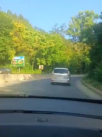 The video shows a view from inside a car driving on a road that curves to the right. The road is bordered by a concrete barrier on the left and a lush green forest on both sides. Several cars are visible on the road. A silver minivan is in front of the camera car, and other cars are further ahead and on the opposite side of the road. A yellow road sign with a curved arrow indicating a sharp turn is visible on the left. In the background, a stone bridge or aqueduct is visible arching over the road. The sky is blue and clear. The lighting suggests it is daytime.
