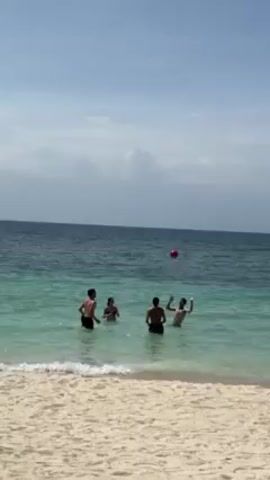 On a sunny day, four shirtless individuals are playing with a red ball in the shallow, clear turquoise water of a beach. The beach has light sand, and the ocean stretches to the horizon under a partly cloudy sky. The individuals are spaced out in the water, actively engaged in tossing the ball. One of them is captured in the midst of jumping to hit the ball with their hands, while others are either preparing to catch or retrieve it.