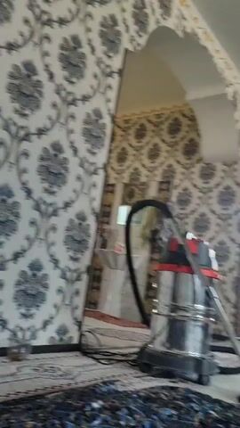 The video shows an interior view of a room with patterned wallpaper. In the foreground, a rug is visible on the floor. Towards the right side of the frame, a metal object, possibly a coffee pot or a kettle, sits on the floor next to a rug. Further into the room, there is a mirror, a chair with striped cushions, and a table with chairs. An arched doorway leads to another area of the room, where a window and a radiator are visible. The room appears to be a dining area or a living space.