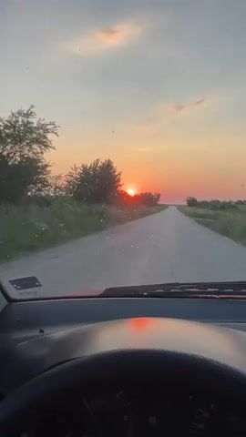 A video shows the view from inside a car looking out the windshield at a road stretching into the distance. The sun is setting, casting an orange and pink glow across the sky. Trees and tall grass line the sides of the road. The dashboard and steering wheel are visible at the bottom of the frame.