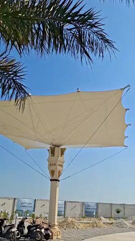 The video pans from a blue sky with palm fronds in the foreground to a black cube-shaped structure with gold accents, resembling the Kaaba. The structure has pillars and an awning extending from its side. The camera then moves to reveal a wider view of an outdoor area. A large white canopy stretches across the sky, supported by poles. In the center of the frame, a small, ornate golden structure on a pedestal is visible, reminiscent of a minaret or a decorative Islamic monument. A wall with trees and construction materials can be seen in the background. The ground is paved with concrete. The overall scene depicts an open, outdoor space with Islamic architectural elements under a clear blue sky.