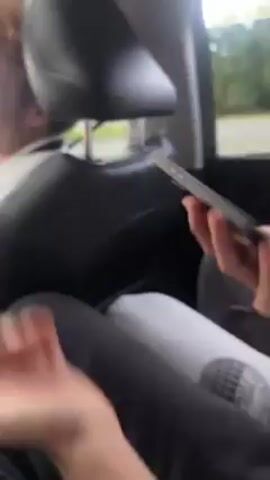 The video shows a person sitting in the backseat of a car, scrolling through messages on their phone. The phone screen displays a messaging interface with various contacts. Another person in the car with red hair turns around and laughs at the camera. The background shows a blurred view of the outside through the car window. The video appears to be taken during a car ride.