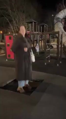 At night, a woman in a long dark puffer coat, light blue jeans, and beige boots jumps on a square-shaped in-ground trampoline in a playground. She holds a white purse over her left shoulder and a clear water bottle in her right hand. She jumps several times, turning around once. The playground features red and gray play structures with ladders and slides. Bare trees are visible in the background. The playground is lit by streetlights.