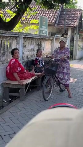 The video shows three women outdoors on a paved street. One woman in a red dress sits on a wooden bench to the left, with her left leg extended. Another woman, in a floral-patterned top, sits beside her. A third woman, wearing a purple and white patterned dress, stands to the right of the bench, holding the handlebars of a bicycle. The bicycle has a black basket on the front. In the background, there is a building with a tiled roof and a sign. Trees and foliage are visible. The lighting suggests it is daytime.