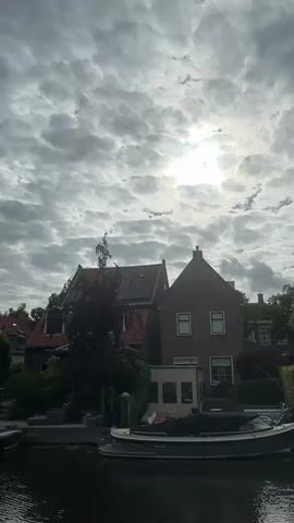 A video displays a cloudy sky with the sun shining through, positioned slightly left of center. Below the sky, a row of houses with red tiled roofs and brown facades are visible. Some houses have solar panels. In front of the houses, a canal runs horizontally across the frame. A boat with a dark cover is docked in the canal. Trees and greenery are interspersed between the houses and along the canal bank. The water in the canal is dark and reflects the scene above. The overall scene depicts a waterside residential area with a boat docked.