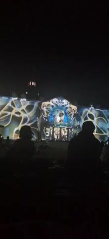A nighttime exterior shot shows a building facade illuminated by a light projection show. In the foreground, the dark silhouettes of several people are seated on the ground, facing away from the camera and towards the building. The building itself is bathed in a dynamic projection of abstract, flowing patterns in shades of yellow, white, and blue. The projection creates an illusion of movement and depth on the architectural surface. The sky above is dark, indicating it is night. The overall scene appears to be an outdoor event or festival.
