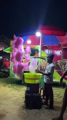 This media file captures a nighttime scene at a street fair or market in Habaraduwa, Sri Lanka. The central focus is a brightly lit candy floss (cotton candy) stall. The stall is adorned with a colorful canopy, a mix of red and blue, and illuminated by overhead lights, creating a vibrant and festive atmosphere.

The primary subjects are two young men, likely vendors or assistants, actively preparing candy floss. One man, wearing a patterned shirt and a cap, is in the process of spinning the candy floss, holding a stick with the pink, sugary creation extending from a machine into a large, bright yellow basin. Another young man, in a dark shirt and brown shorts, stands nearby, also holding a bag, possibly to package the candy floss. In the background, a younger boy can be seen through the stall's window, also engaged with the candy floss machine.

Hanging prominently from the stall are several bags of pre-made pink candy floss, showcasing the product. The stall itself appears to be on wheels, suggesting it's a mobile vendor. The background reveals a busy fairground setting with other stalls, a motorcycle, and people dressed in casual attire. The ground is sandy or dusty, typical of an outdoor event.

The time of day is clearly night, indicated by the darkness and the artificial lighting. There are no immediate cues about the weather, but the dry ground suggests it's not raining. The overall mood conveyed is lively and energetic, typical of a nighttime market or fair.

Visible text includes what appears to be a price or product information on the stall's side, partially obscured by the canopy and the people. The visual context strongly suggests a commercial activity focused on selling sweet treats at a local event in Sri Lanka.