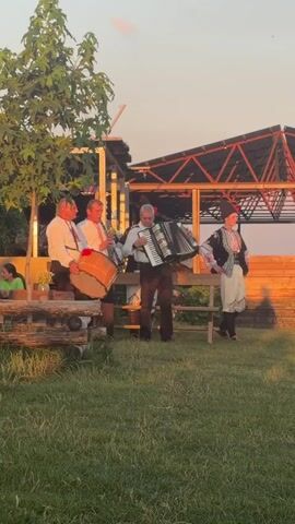 A group of people are playing music and dancing outdoors at sunset. In the foreground, a man plays an accordion while two other men play drums. To the right, a person in traditional clothing stands with their hands on their hips. In the background, several women in traditional clothing are dancing in a circle. The scene appears to be set in a rural area with a grassy field and a wooden structure. The lighting suggests it is late afternoon or early evening.