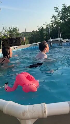 The video shows two children swimming in an above-ground pool. A girl with dark hair is on the left, and a boy with dark hair is on the right. Both children are in the water, with the boy appearing to splash or move around more actively. There are several inflatable pink flamingos floating in the pool, as well as a blue inflatable ring. The pool has a silver metal ladder with white steps visible at the far end. The background shows green trees and a clear sky with the sun visible, suggesting a daytime setting. The poolside is partially visible in the foreground, with white pipes and a grey edge. The girl is facing the boy, and they appear to be interacting, possibly playing. The boy is also looking towards the girl at one point. The water is blue and rippling.