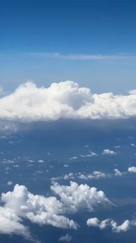 The video shows a view from an airplane looking down at clouds and the sky. The sky is a bright blue with some thin, wispy clouds higher up. Below, there are large, fluffy cumulus clouds and smaller, scattered clouds. The ground is visible through gaps in the clouds, appearing as a dark, possibly mountainous landscape. The overall scene is one of aerial perspective above a cloud-covered landscape under a clear blue sky.