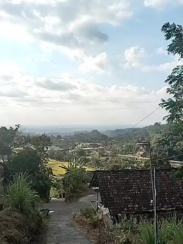 A high-angle shot looks out over a valley dotted with houses and trees. A road winds down into the valley from the foreground. The sky is cloudy, and the sun is shining. The scene is peaceful and serene.