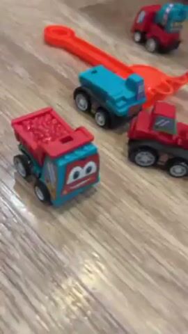 The video opens with a close-up of a light wood floor and several toy construction vehicles scattered around. Some of the vehicles are red and blue, while others are red and light blue. An orange toy track is also visible. The camera moves slightly to show a yellow and blue toy excavator. The camera then focuses on a blue tray filled with gray kinetic sand. A pair of hands, with manicured fingernails and a ring on one finger, come into the frame and begin packing the sand into an orange mold. The mold is then turned over, releasing a sand sculpture.