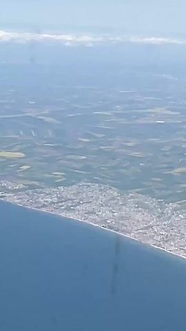 The video shows an aerial view from an airplane, looking out the window. The landscape below is a patchwork of fields, with a dense urban area along the coastline. The sea is a deep blue. A red and white Turkish Airlines wing is visible in the corner of the frame. The sky is clear blue with some distant clouds on the horizon.
