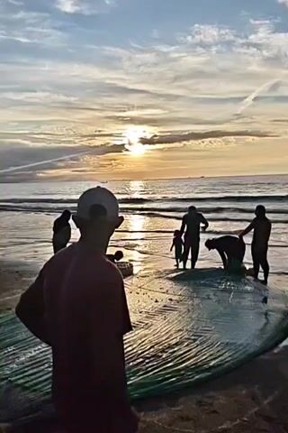 A group of silhouettes are on a sandy beach at sunset. The sun is low in the sky, casting a warm glow over the scene. In the foreground, a person wearing a red shirt and a white cap stands with their hands on their hips, facing away from the camera and towards the ocean. In the middle ground, several other figures are gathered around a large, green net spread out on the sand. Some individuals appear to be working with the net, while others are standing and observing. The ocean is visible in the background, with gentle waves lapping at the shore. The sky is filled with dramatic clouds, illuminated by the setting sun. The overall impression is one of a coastal community engaged in fishing activities at the end of the day.
