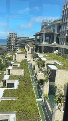 The camera pans across a high-angle shot of a tropical resort with modern architecture. The foreground is dominated by buildings with green roofs and narrow, rectangular pools. In the middle ground, a cluster of buildings is surrounded by lush greenery, including palm trees. The background reveals a vast ocean under a bright blue sky with scattered clouds. The scene is viewed through a window, with reflections visible on the glass. The overall atmosphere is serene and tropical.