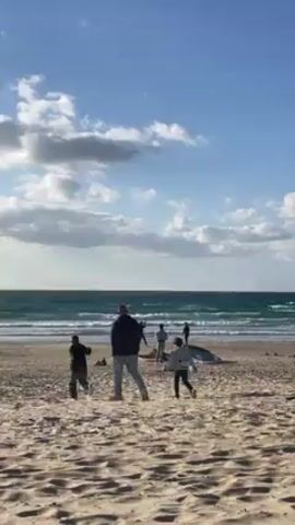 The video shows a beach scene on a partly cloudy day. Several people are visible on the beach. A man and two children walk towards the water. Other people are standing near surfboards lying on the sand. The ocean has waves.