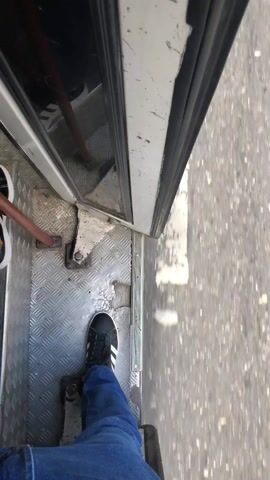 The video captures a first-person, downward-facing view of a person's legs and feet at the open doorway of a moving vehicle. The person is wearing blue jeans and black Adidas sneakers with white stripes. One foot is positioned on a metallic, diamond-plate textured floor inside the vehicle, near the open door. A white door frame is visible on the left, along with a metal bar or handle. Outside the vehicle, the ground, appearing to be an asphalt road with a faint white line, is blurred, indicating that the vehicle is in motion. The bright lighting suggests it is daytime.