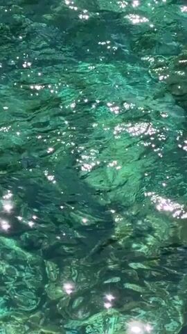 The video starts with a close-up view of clear turquoise water with sunlight glinting off the surface. The camera then pans up to reveal a coastal scene with rocky cliffs on either side of a small bay. The water is exceptionally clear, showing the rocky bottom beneath. In the distance, the ocean extends to the horizon under a clear blue sky, with a few boats visible. Vegetation grows on the clifftop. The camera remains static for the rest of the video, presenting the picturesque view.