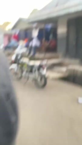 The video presents a shaky, handheld horizontal pan shot across a street scene in what appears to be a village or town. Multiple single-story buildings with corrugated metal roofs and painted walls (pink, gray, brick) line the dirt road. Numerous people are visible, primarily sitting in groups on steps, benches, or stools outside the buildings. Several motorcycles of various colors (white, yellow, red, black) are parked along the roadside. A dark-colored sedan is also briefly seen parked. Signs are visible on some of the buildings. Trees and hills can be seen in the background. The general activity involves people observing and resting.