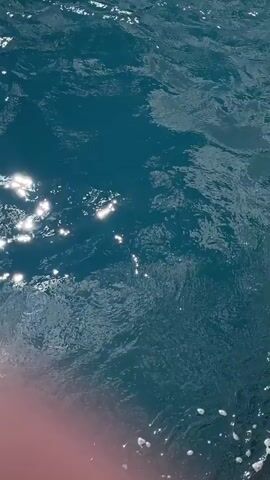 The video shows a close-up view of dark blue water. Sunlight glints off the surface, creating shimmering highlights. White foam is visible on the left and lower right sides of the frame, suggesting movement or disturbance of the water. The bottom left corner shows a sliver of a pink object, possibly part of a boat or personal flotation device. The water appears somewhat choppy.