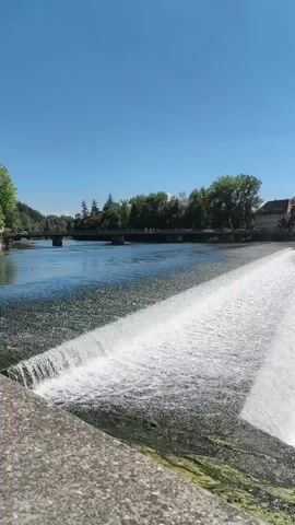 The video shows a view of a waterfall in a river. The sky is clear and blue, and the sun is shining. There are trees and buildings in the background. The water is flowing over a concrete dam, creating white foam. The camera is positioned on a stone wall overlooking the waterfall. The video is taken from a stationary position, and there is no visible movement of the camera. The overall impression is one of a peaceful and scenic natural environment.