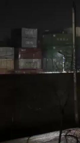 The video shows a night scene featuring a stack of shipping containers. The containers are various colors, with visible labels such as "CHIYU" and "MARITIME" on some. In the background, there's a blurred figure standing near a light source. The foreground is dark, with a wall or barrier partially obscuring the view and a bare tree branch visible on the right. Utility poles and wires are also visible.
