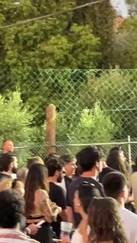 The video shows a crowd of people standing behind a fence and a metal barrier. They appear to be at an outdoor event, possibly a concert or festival, with trees and lighting rigs visible in the background. In the foreground, several people are facing away from the camera, looking towards a stage. On the stage, a person is playing a guitar and singing into a microphone. There are large speakers and amplifier equipment on the stage. The lighting is dim, suggesting it is evening or nighttime.