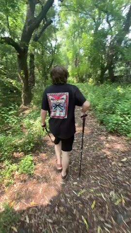 A person walks away from the camera on a dirt trail in a forest. The person is wearing a black t-shirt with a colorful design on the back, black shorts, and brown shoes. They are using trekking poles to help them walk. The trail is surrounded by trees and green plants.